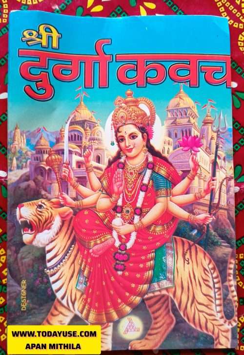 A full-color devotional image on a book cover. The central figure is the Hindu goddess Durga, depicted with multiple arms, riding a tiger. She holds various weapons and symbols, including a sword, trident, and lotus flower. She wears a crown, a red sari, and elaborate jewelry. In the background, several ornate temples or palaces with golden domes are visible against a blue sky. At the top of the image, the title 'श्री दुर्गा कवच' (Shri Durga Kavach) is written in large red Hindi script with a yellow outline, above the illustration. A website URL, 'WWW.TODAYU56.COM', is partially visible at the bottom left, and 'DESIGNER' is written vertically on the left side." Apan Mithila www.todayuse.com
